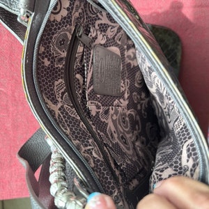 Op de afbeelding: Een grijze leren handtas met een zwart-witte bloemenvoering. De tas heeft een ritsvak en een zilveren kettingband.