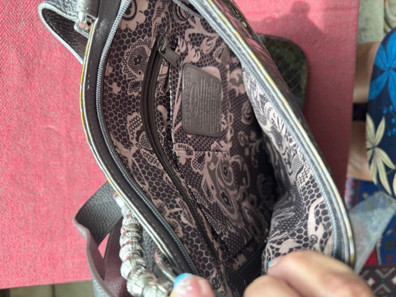 Op de afbeelding: Een grijze leren handtas met een zwart-witte bloemenvoering. De tas heeft een ritsvak en een zilveren kettingband.