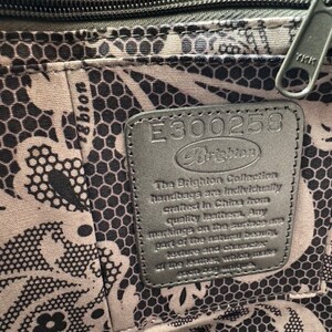 Op de afbeelding: Close-up van een zwart lederen label met de tekst "E300258 Brighton" en een beschrijving van de Brighton Collection handtassen. Het label is bevestigd aan een zwart-wit gepatroneerd weefsel.