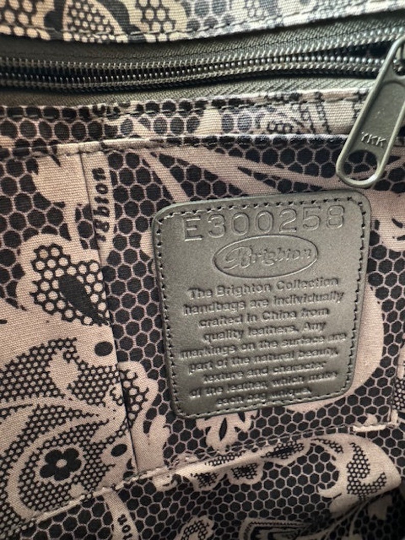 Op de afbeelding: Close-up van een zwart lederen label met de tekst "E300258 Brighton" en een beschrijving van de Brighton Collection handtassen. Het label is bevestigd aan een zwart-wit gepatroneerd weefsel.
