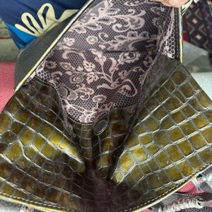 Op de afbeelding: Een groene handtas van krokodilleer met een zwarte voering en een bloemenpatroon. De tas heeft een bovenhandvat en een schouderband.