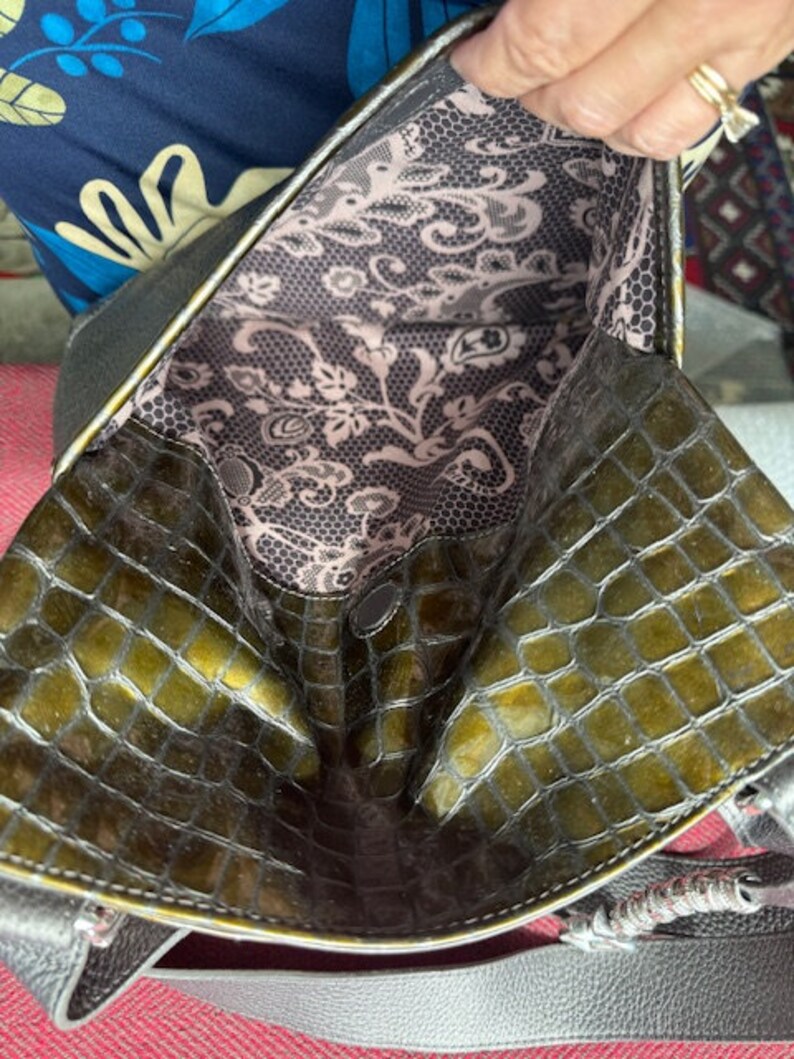 Op de afbeelding: Een groene handtas van krokodilleer met een zwarte voering en een bloemenpatroon. De tas heeft een bovenhandvat en een schouderband.