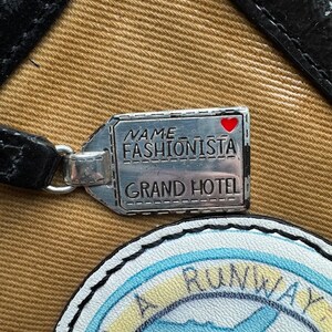 Puede incluir: Una etiqueta de equipaje plateada con el texto "NAME FASHIONISTA GRAND HOTEL" y un coraz&oacute;n rojo. La etiqueta est&aacute; unida a una bolsa de lona marr&oacute;n con un parche de avi&oacute;n azul y blanco que dice "RUNWAY".