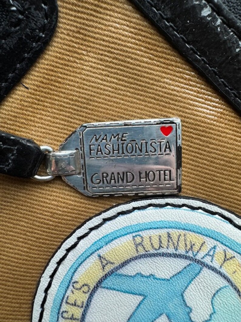 Puede incluir: Una etiqueta de equipaje plateada con el texto "NAME FASHIONISTA GRAND HOTEL" y un coraz&oacute;n rojo. La etiqueta est&aacute; unida a una bolsa de lona marr&oacute;n con un parche de avi&oacute;n azul y blanco que dice "RUNWAY".
