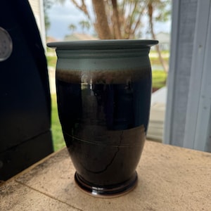 May include: A handmade ceramic vase with a dark blue, brown, and teal glaze. The vase has a cylindrical shape with a slightly flared rim and a rounded base. The exterior has a glossy finish.