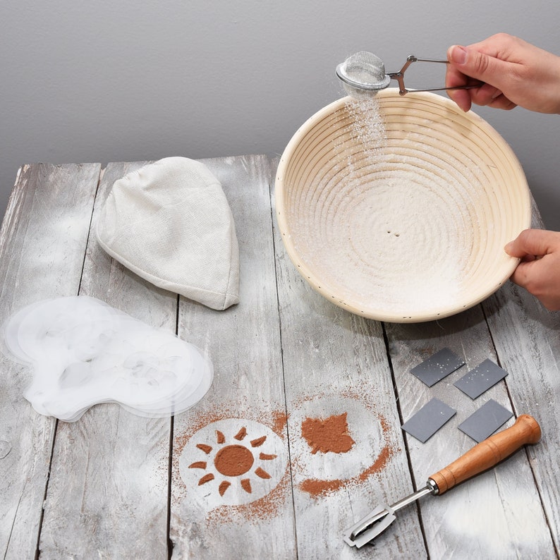 hand holding flour sifter sifting flour into banneton basket and sun/ leaf shaped pattern on table shaped by using bread stencils cloth linen and bread lame on the side on a grey table