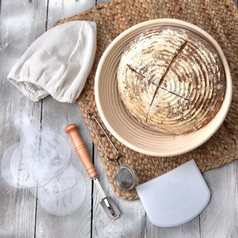 artisan bread in proofing basket on a mat with accessories including linen cloth bread lame dough scraper flour sifter stencil set