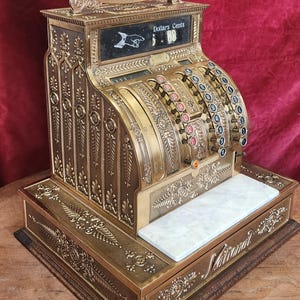 May include: Antique brass cash register with ornate floral detailing. The register displays "Amount Purchased" and "Dollars Cents" with a digital display. Features a white marble shelf and a wooden base. The word "National" is visible.