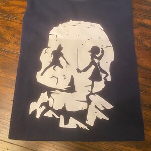 Puede incluir: Una camiseta azul marino con una silueta blanca de dos figuras de pie frente a una calavera.