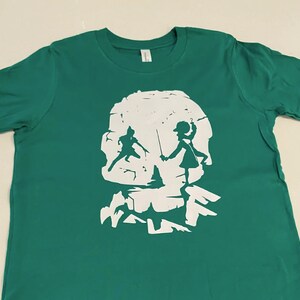 Puede incluir: Una camiseta verde con una silueta blanca de un cráneo. Dentro del cráneo hay dos figuras, una sosteniendo una espada y la otra un bastón. Las figuras se enfrentan.