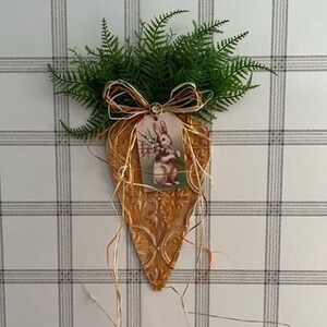 May include: A brown and gold carrot-shaped wall decor with a vintage bunny illustration and a brown bow. The decor is hanging on a white wall with a gray and white plaid pattern.