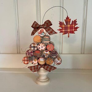 Peut inclure: Un arbre décoratif sur le thème de l'automne, fait de bouchons, orné de papier à motifs et d'un nœud écossais. L'arbre est posé sur un socle en bois tourné blanc. Une feuille à carreaux est suspendue au-dessus de l'arbre.