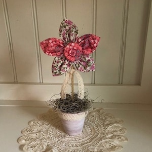 Puede incluir: Una flor de tela rosa y blanca con un botón en el centro en una maceta blanca pequeña con un encaje debajo.