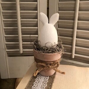 Puede incluir: Un huevo de conejo blanco en forma de conejo en una pequeña maceta de terracota con cuerda marrón y cinta de arpillera. La maceta está sobre una superficie marrón claro.