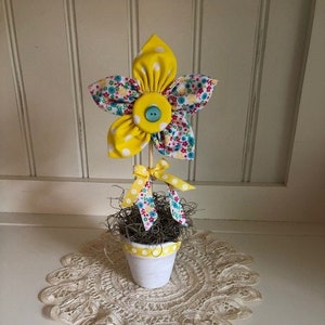 Puede incluir: Una flor de tela amarilla y blanca con un botón azul en el centro en una maceta blanca con lunares amarillos. La maceta está sobre un encaje blanco de ganchillo.