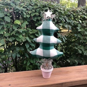 Puede incluir: Un pequeño árbol de Navidad a cuadros verde y blanco con una estrella blanca en la parte superior, sentado en una maceta rosa. El árbol está en una terraza de madera.