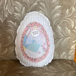 Puede incluir: Almohada blanca de ganchillo en forma de huevo con un diseño de patchwork de tela rosa y blanca en el interior. La tela tiene un patrón floral y el texto "Spring Wishes" es visible.