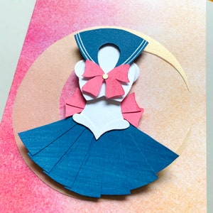 May include: A papercraft Sailor Moon costume, featuring a navy blue sailor collar and skirt, a white top, and pink bow accents. The design is set against a pink and peach watercolor background, with a crescent moon shape.