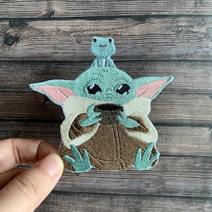 Op de afbeelding: Geborduurde patch met een groen, buitenaards personage dat een beker vasthoudt, met een kleine kikker op zijn hoofd. De patch heeft een lichtgroen, bruin en beige kleurenschema met roze accenten op de oren.