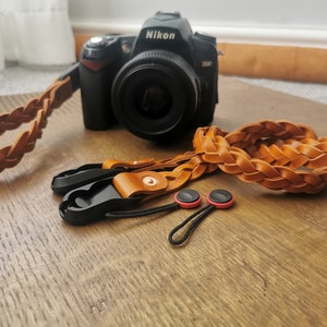 May include: A Nikon D90 camera with a brown, plaited leather wrist strap and a matching camera strap. Also included are two black and brown camera connectors with red accents.