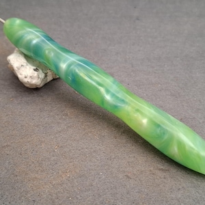 Può includere: Un uncinetto con un manico ergonomico e liscio caratterizzato da un motivo marmorizzato nei toni del verde chiaro, lime e verde acqua. Il manico ha una forma affusolata e sagomata per la presa, con un sottile gancio in metallo argentato.
