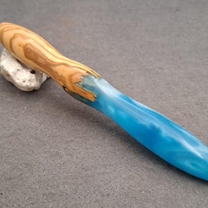 Op de afbeelding: Een haaknaald met een handvat gemaakt van een combinatie van lichtbruin hout met een natuurlijk nerfpatroon en een glad, doorschijnend blauw harsgedeelte dat taps toeloopt naar het uiteinde, bevestigd aan een dunne metalen haakpunt.