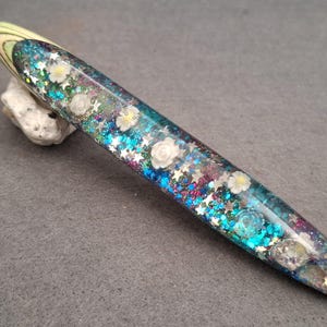 Op de afbeelding: Een haaknaald met een taps toelopend handvat van heldere hars, ingelegd met blauwe, paarse en turquoise glitters, kleine zilveren stervormige lovertjes en diverse kleine witte bloemvormige versieringen, met een lichtgroene en crèmekleurige top.