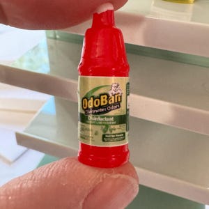 Puede incluir: Una peque&ntilde;a botella roja de desinfectante OdoBan, sostenida entre los dedos. La etiqueta es verde y dorada, con el nombre del producto y las palabras "Elimina olores" y "Desinfectante" visibles. La botella es peque&ntilde;a, probablemente para una casa de mu&ntilde;ecas.