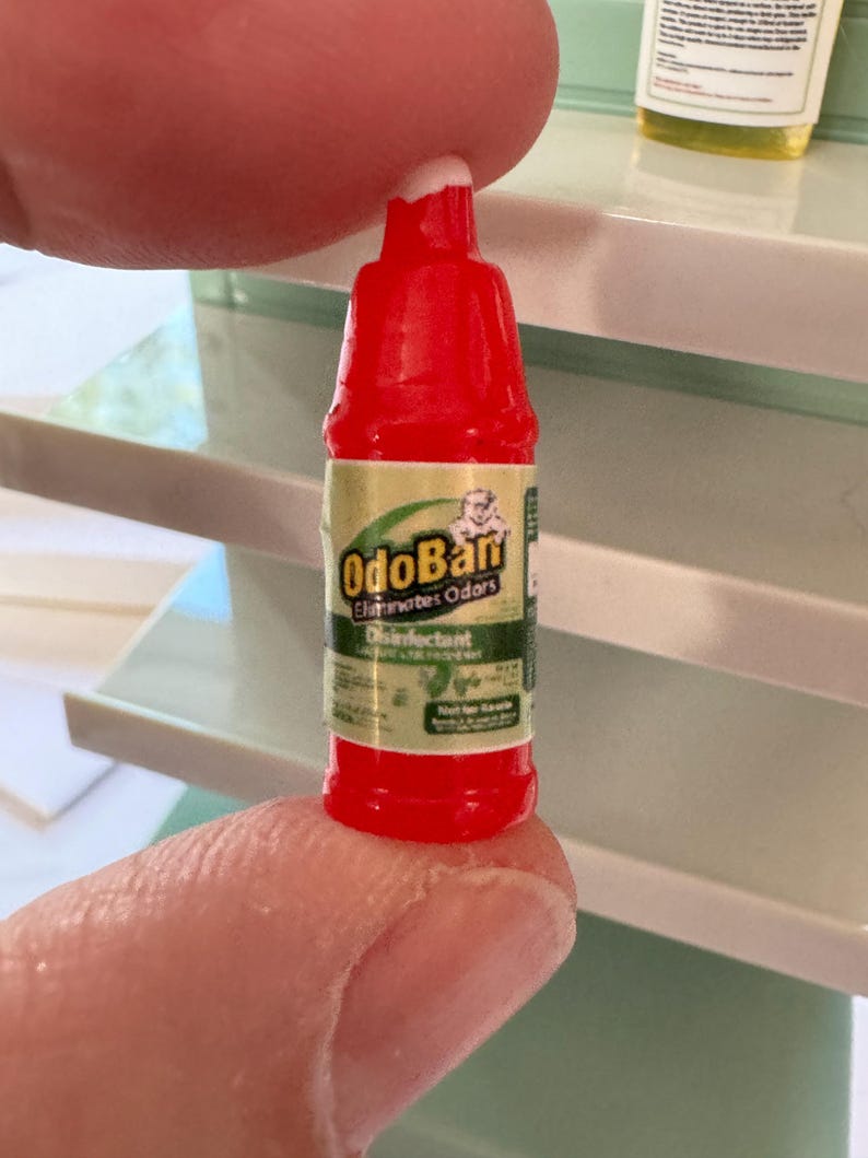 Puede incluir: Una peque&ntilde;a botella roja de desinfectante OdoBan, sostenida entre los dedos. La etiqueta es verde y dorada, con el nombre del producto y las palabras "Elimina olores" y "Desinfectante" visibles. La botella es peque&ntilde;a, probablemente para una casa de mu&ntilde;ecas.