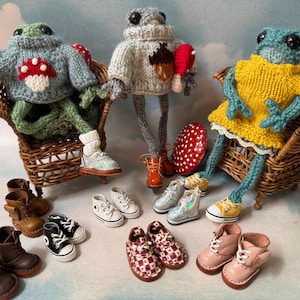May include: Handmade frog dolls wearing sweaters and dresses, seated in miniature wicker chairs. Various pairs of tiny shoes are arranged in front of the dolls, including boots, sneakers, and dress shoes. A whimsical, handcrafted theme.