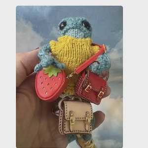 May include: A handmade crocheted turtle doll wearing a yellow sweater and carrying three miniature bags: a red strawberry-shaped purse, a red satchel, and a tan backpack. The turtle is blue and has black eyes.