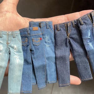 May include: A collection of miniature denim jeans in various shades of blue, held in a hand. The jeans have classic details like pockets, belt loops, and stitching. Some jeans have distressed detailing.