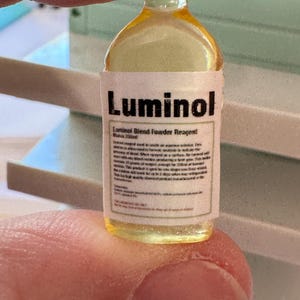 Puede incluir: Una peque&ntilde;a botella transparente con un l&iacute;quido amarillo, etiquetada como "Luminol Blend Powder Reagent". La etiqueta incluye texto adicional. La botella se sostiene entre los dedos.