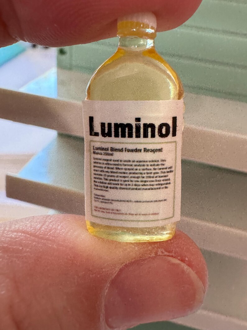 Puede incluir: Una peque&ntilde;a botella transparente con un l&iacute;quido amarillo, etiquetada como "Luminol Blend Powder Reagent". La etiqueta incluye texto adicional. La botella se sostiene entre los dedos.