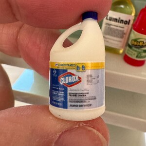 Puede incluir: Botella de lej&iacute;a Clorox en miniatura con tapa azul, cuerpo blanco y una etiqueta con detalles en azul, amarillo y rojo. La botella se sostiene entre los dedos, con otras botellas en miniatura en el fondo.
