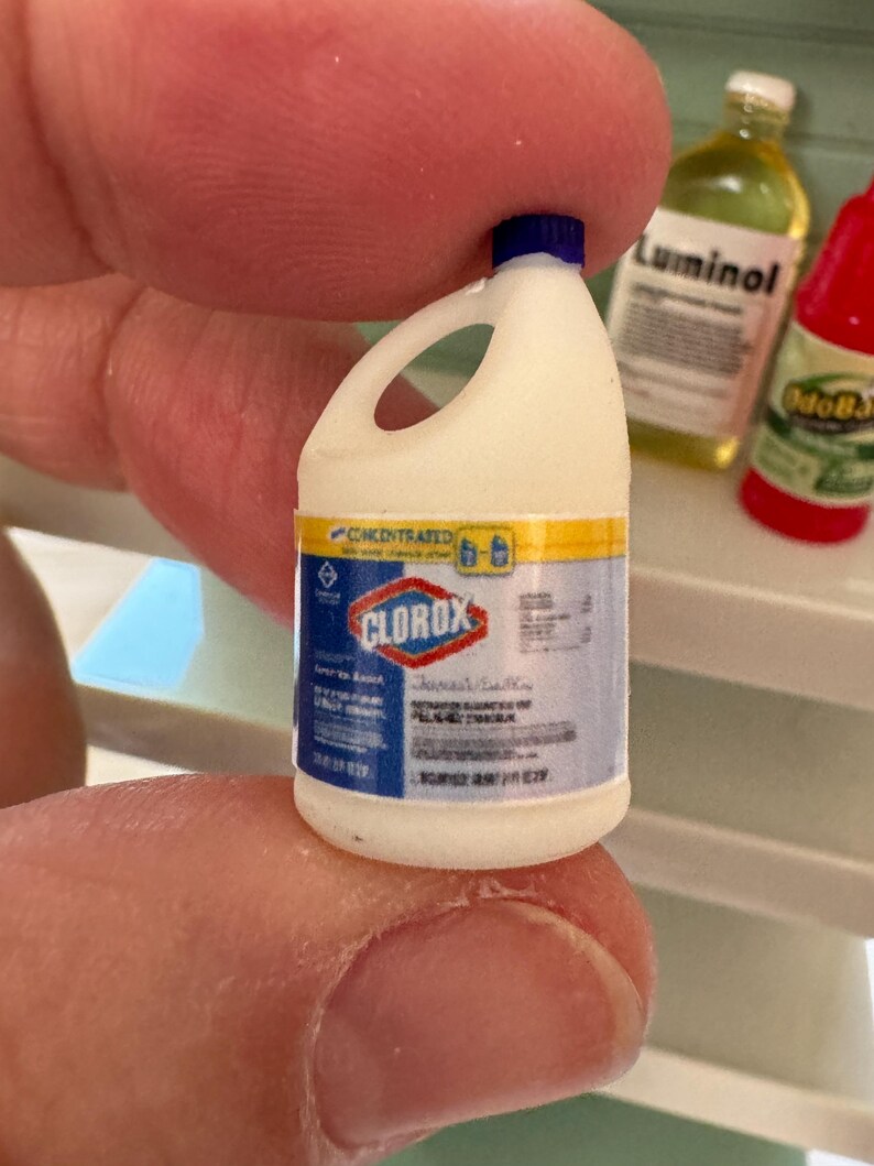 Puede incluir: Botella de lej&iacute;a Clorox en miniatura con tapa azul, cuerpo blanco y una etiqueta con detalles en azul, amarillo y rojo. La botella se sostiene entre los dedos, con otras botellas en miniatura en el fondo.