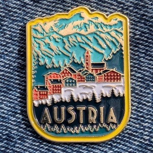 May include: A gold-colored enamel pin with a blue and green mountain scene and a small village in the foreground. The text "AUSTRIA" is written in gold letters on the bottom of the pin.