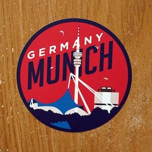 Peut inclure: Un autocollant circulaire rouge et bleu avec le texte "GERMANY MUNICH" et une illustration stylisée d'une tour et d'un bâtiment.