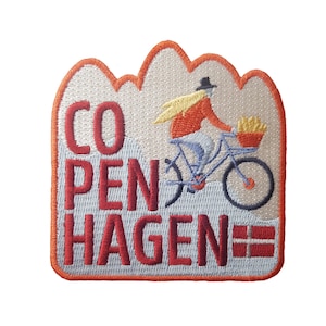 以下が含まれることがあります： 白い背景とオレンジ色の縁取りのある刺繍パッチ。パッチには、赤、白、青のフランスフライの入ったバスケットを持った自転車に乗っている人のイラストが描かれています。パッチには赤い糸で「COPENHAGEN」という文字が刺繍されています。