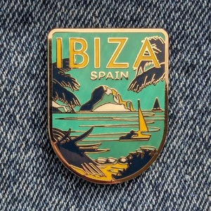 Könnte beinhalten: Goldfarbene Emaille-Anstecknadel mit einem grün-blauen Design. Die Anstecknadel zeigt ein Segelboot, eine kleine Insel und Palmen. Der Text "IBIZA SPANIEN" ist auf der Anstecknadel aufgedruckt.