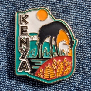 Può includere: Uno spillo in smalto dorato con un designo verde, bianco e arancione. Lo spillo presenta una giraffa nera e bianca e un vitello bianco, insieme alla parola "KENYA" in lettere nere.