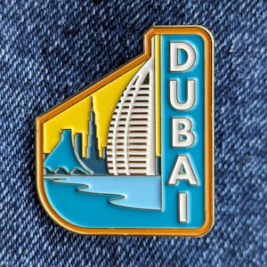 May include: A blue and yellow enamel pin with a white building and the word "DUBAI" in white letters.
