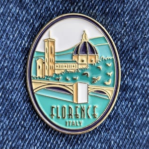 May include: Gold enamel pin with a white background depicting a cityscape of Florence, Italy. The pin features a bridge, a building with a dome, and trees. The text "FLORENCE ITALY" is printed below the image.