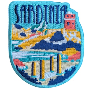 Puede incluir: Un parche bordado azul y blanco con la palabra "SARDINIA" en letras azules. El parche presenta una ilustración colorida de una escena de playa con un faro, una playa de arena y agua azul.