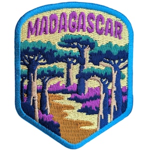 Puede incluir: Un parche bordado con un borde azul y un fondo amarillo. El parche presenta una imagen estilizada de árboles y un camino en tonos de morado, verde y marrón. El texto "MADAGASCAR" está bordado en letras moradas en la parte superior.