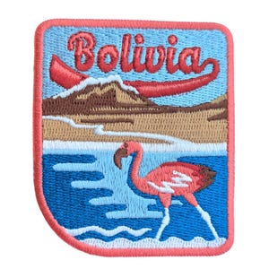 May include: Embroidered patch with a blue, white, and brown landscape scene with a red boat and the text "Bolivia". A pink flamingo stands in the water.