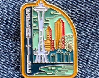 Peut inclure: Une épingle rectangulaire en émail représentant le paysage urbain de Seattle. L'épingle comprend la Space Needle, des gratte-ciel et une grande roue, avec le mot "SEATTLE" en texte vertical. L'épingle a une bordure dorée et est posée sur un fond en denim bleu.