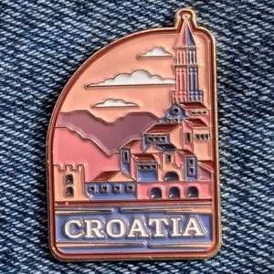 Op de afbeelding: Een gouden emaille pin met een roze en paarse illustratie van een skyline van een stad met een hoge toren. De pin heeft het woord "CROATIA" in gouden letters op een blauwe achtergrond.