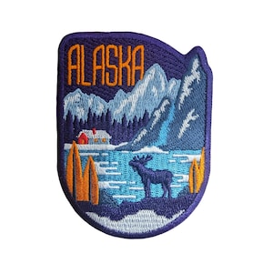 以下が含まれることがあります： 青い背景に山々の風景が刺繍されたパッチ。パッチの上部にはオレンジ色の文字で「アラスカ」と書いてあります。遠くには小さな小屋が見え、手前にはヘラジカが立っています。