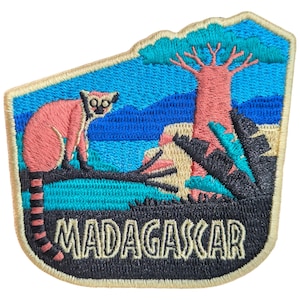 May include: Embroidered patch depicting a pink lemur on a branch, with a pink tree and the word "MADAGASCAR". The background shows blue water and sky, with a black and teal foreground. The patch has a cream-coloured border.